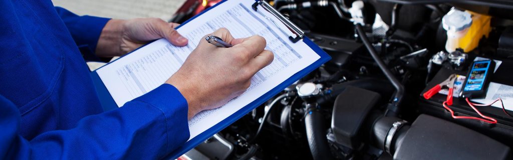 A mechanic filling out a checklist on a clipboard over an open car engine bay.